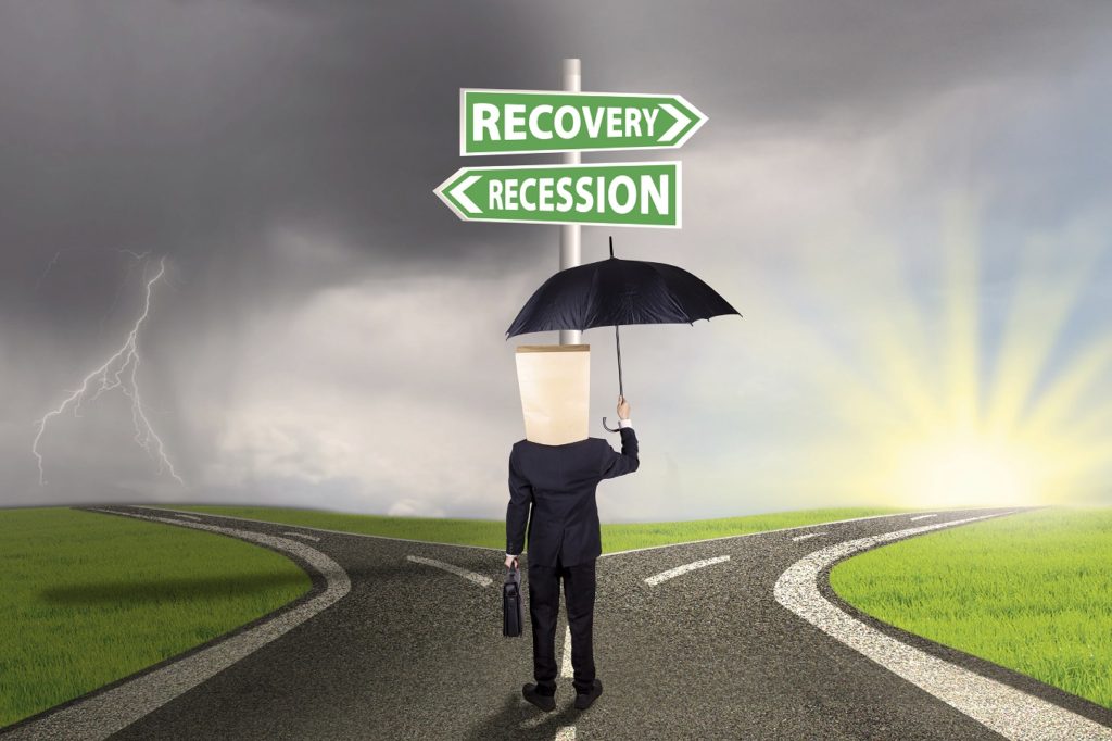 Worker with cardboard head and umbrella, standing on the road while looking at signpost directing at the road to recovery or recession financial